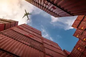 Blick zwischen aufeinander gestapelten roten Containern nach oben auf den Himmel auf ein Flugzeug.