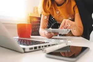 Frau sitzt mit einem Tablet in der Hand am Tisch. Vor ihr sind Tasse, Notebook, Kugelschreiber und Smartphone. Kleine Icons sind über dem Tablet miteinander verbunden.