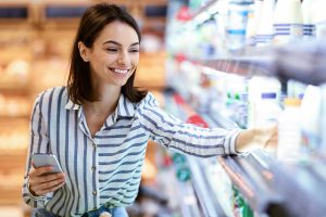 Eine Frau greift in einem Supermarkt zu einer Milchflasche und hält ein Smartphone in der Hand.