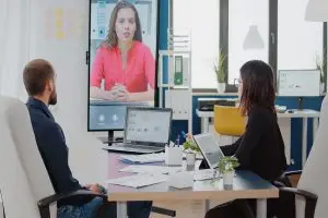 Ein Mann und eine Frau sitzen im Büro am Schreibtisch und unterhalten sich per Videokonferenz mit einer Kollegin.