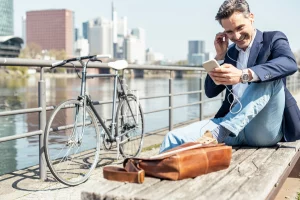 Ein Mann im Sakko sitzt an der Spree und schaut auf sein Handy